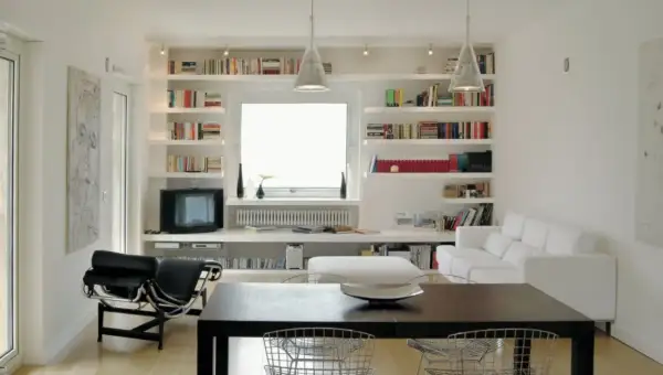 Soggiorno moderno con ampie librerie cartongesso a parete, piene di libri, che incorniciano una finestra. Divano bianco e poltrona nera completano l'arredo.