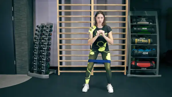 Kobieta w sportowym stroju wykonuje ćwiczenie monster walk z gumą oporową na udach.