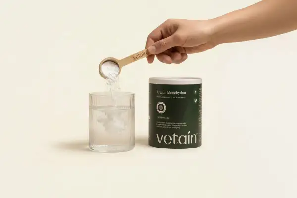 Eine Hand schüttet Kreatinpulver aus einem Löffel in ein Glas Wasser. Das Pulver löst sich auf, was auf die Einnahme von Kreatin hindeutet, das manchmal mit Wassereinlagerung in Verbindung gebracht wird.