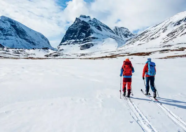 Två skidåkare med ryggsäckar på väg att bestiga berg i snöklädda fjäll under en molnig himmel.