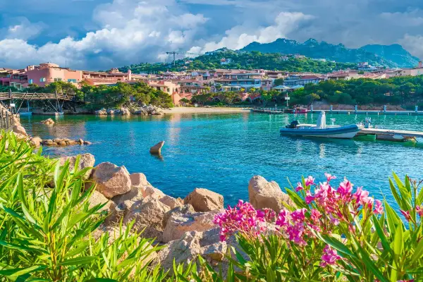 Vue d'une baie turquoise en Sardaigne, avec des maisons roses, un pont en bois et des fleurs roses. Idéal pour savoir que faire en Sardaigne.