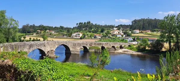 El ponte de Outeiro, un puente de piedra con varios arcos, cruza un río azul. En la orilla opuesta, casas con tejados rojos se asientan en una colina verde.