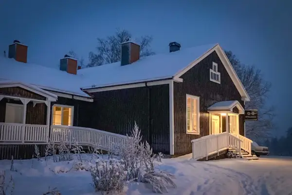 Mörkt trähus med snö på taket och runt om. Varmt ljus lyser från fönster och entré. Skylt med texten "Stilleben" syns.