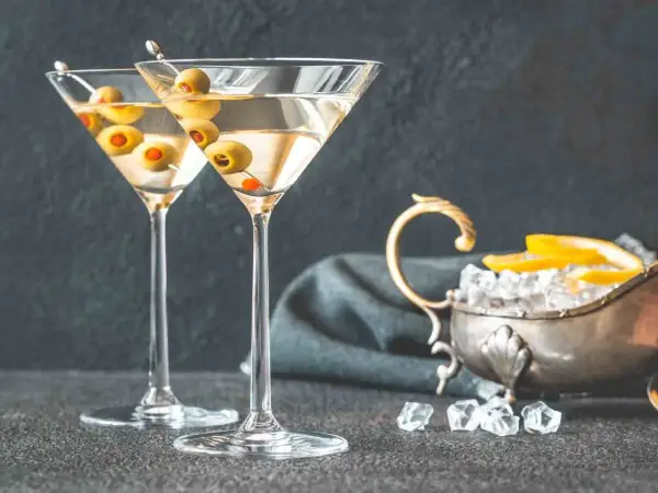 Dwa kieliszki z martini, ozdobione oliwkami, czekają na degustację. Obok lód i cytryna, idealne dodatki, by dowiedzieć się, jak pić martini.