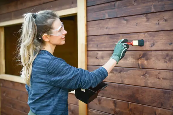 Donna con occhiali protettivi applica vernice su una parete di legno scuro. Si può verniciare sopra l'impregnante per un nuovo look.