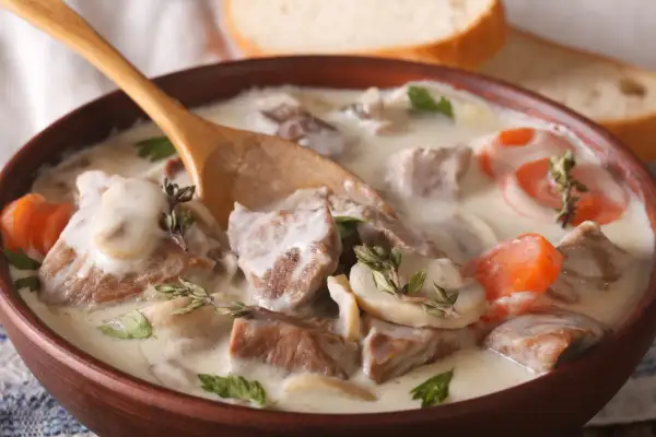 Un plat réconfortant de véritable recette blanquette de veau, avec des morceaux de viande tendre, des champignons et des carottes dans une sauce crémeuse, servi dans un bol en bois avec une cuillère.