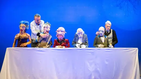 Puppenspieler dirigieren ihre Figuren auf der Bühne im Theater Remscheid. Eine bunte Truppe von Marionetten sitzt hinter einem weißen Tuch.