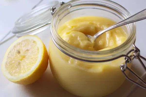 En burk med krämig lemon curd och en halv citron bredvid. En sked vilar i burken, redo att skopa upp den syrliga godsaken.