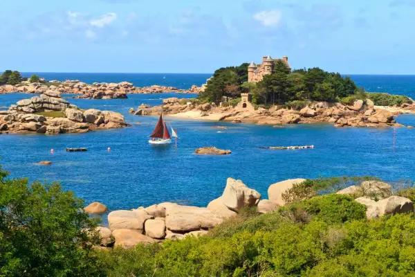 Un voilier navigue près d'un château sur une île rocheuse en Bretagne. Le paysage est typique de la côte bretonne.