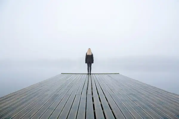 Una persona fredda, con la psicologia che la isola, sta in piedi su un molo di legno in una giornata nebbiosa.