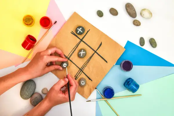 Mani che creano un gioco del tris fai da te dipingendo simboli su sassi. Materiali colorati e pennelli completano la scena.