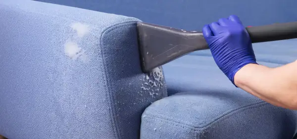 Blaue Polster reinigen mit Rasierschaum. Ein Handschuh hält ein Reinigungsgerät, das Schaum auf den Stoff sprüht.