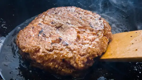 Jugosa carne dorándose en sartén, parte de como hacer hamburguesas caseras. Humo y burbujas de grasa.