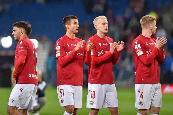 Piłkarze w czerwonych koszulkach i białych spodenkach klaszczą na boisku.