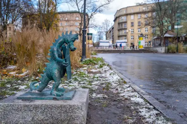 Mały smok z brązu, symbol smoczy szlak kraków, stoi na kamiennym postumencie. W tle miejski krajobraz z budynkami i drzewami.