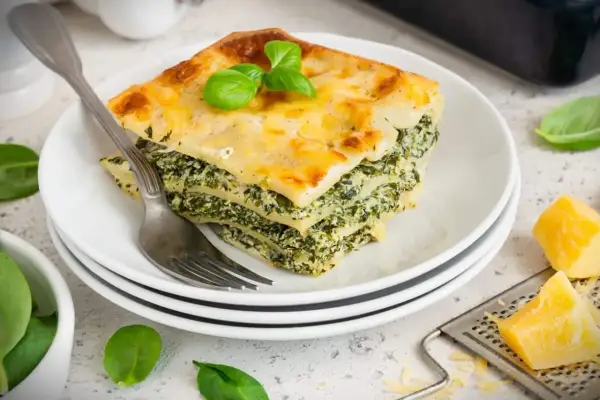 Ein Stück Spinatlasagne mit Käse überbacken, garniert mit Basilikum, auf einem Teller mit einer Gabel.