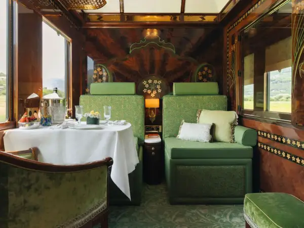 Luxuriöser Speisewagen im Orient Express: Ein gedeckter Tisch mit Obst und Champagner, grüne Sitzmöbel und kunstvolle Holzverzierungen.