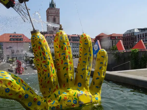 Żółta, kolorowa dłoń fontanny w centrum Frankfurtu nad Odrą, z widoczną wieżą kościelną w tle.