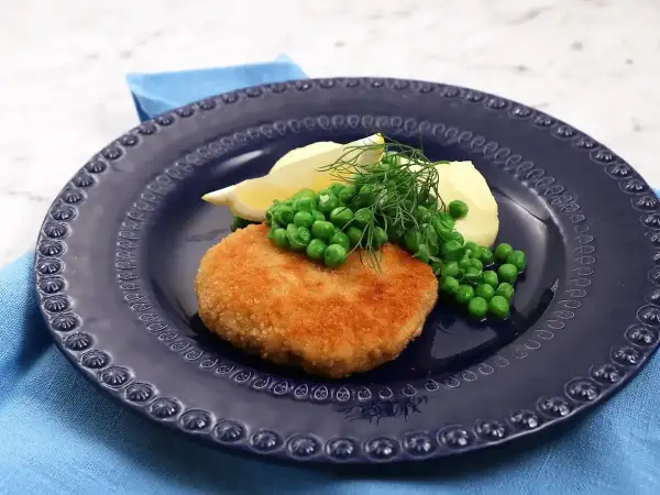 En gyllene havets wallenbergare serveras med potatis, gröna ärtor, citronklyfta och dill på en mörkblå tallrik.