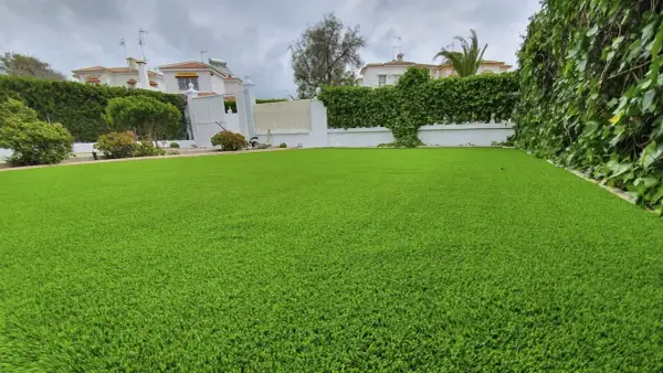 Un jardín con césped artificial impecable, delimitado por vegetación exuberante y casas blancas al fondo.
