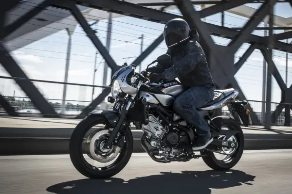 Motocyklista w kasku jedzie srebrnym cafe racerem Suzuki SV650X po moście.