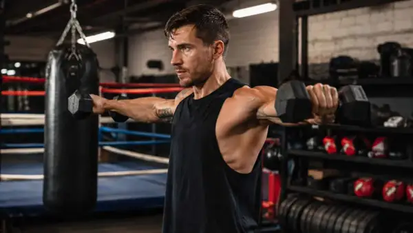 Man performing a boxing shoulder workout with dumbbells in a gym. He's focused, with toned arms and shoulders.