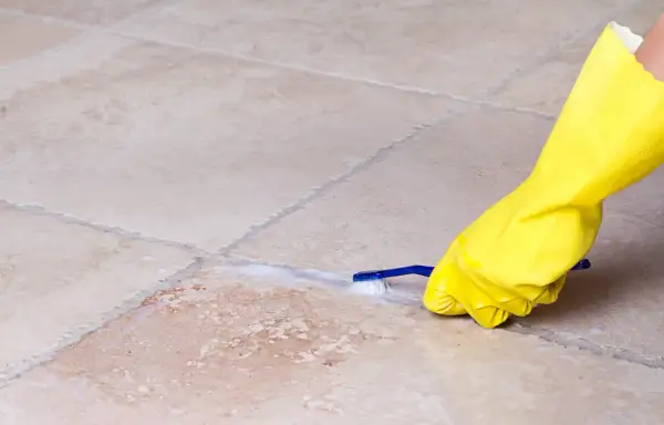 Mano con guanto giallo pulisce le fughe dei pavimenti con uno spazzolino e schiuma.