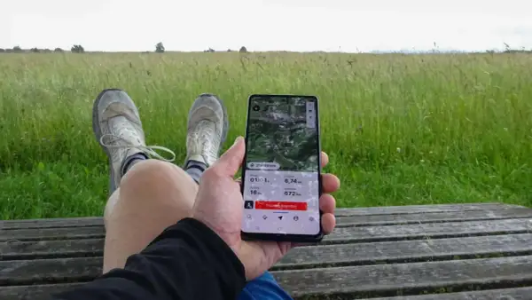 Person sitzt auf einer Holzbank, hält ein Smartphone mit einer guten Wander-App, die eine 6,74 km lange Tour mit 16 hm Anstieg und 672 hm Seehöhe anzeigt.
