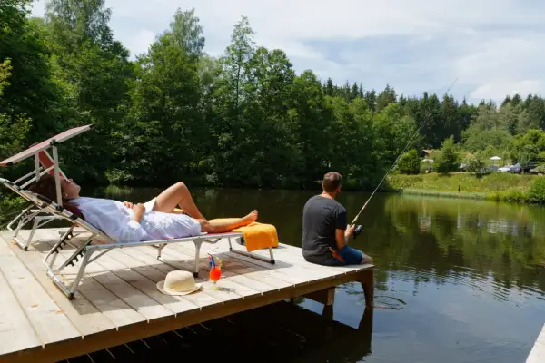 Ein Mann angelt, während eine Frau auf einer Liege entspannt. Perfekter Urlaub in Deutschland am See mit Blick auf den Wald.