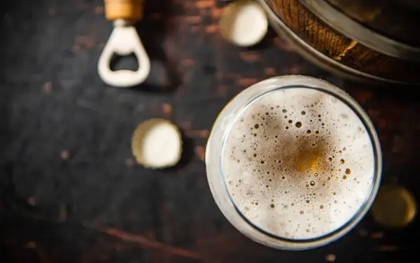 Un bicchiere di birra con una schiuma dorata e bollicine, accanto a un apribottiglie e tappi su uno sfondo scuro. La birra senza schiuma è un piacere per gli occhi.