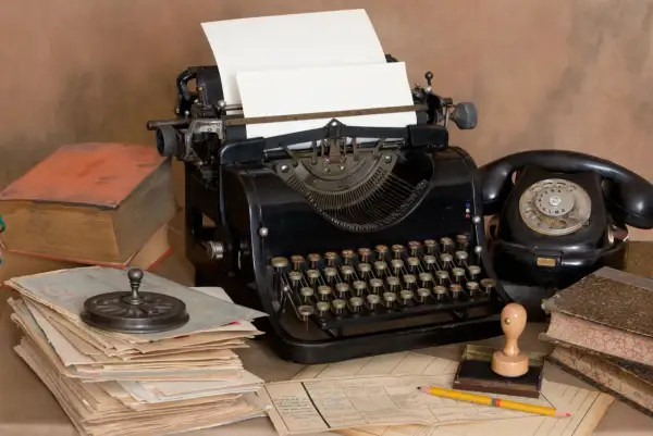 Escritorio antiguo con máquina de escribir, teléfono negro, libros y papeles. Un lápiz amarillo descansa sobre documentos, evocando la creación de dibujos con máquina de escribir.