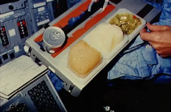 En astronaut äter rymdmat från en bricka. Maten ser ut som en burk, en gelé och en grönsaksrätt.