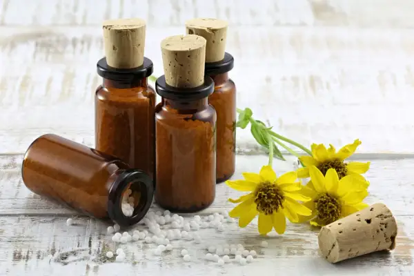Bursztynowe buteleczki z korkami i białymi kuleczkami, symbolizujące homeopatię, obok żółtych kwiatów na drewnianym tle.