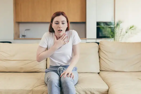 Une jeune femme assise sur un canapé, la main sur la poitrine, le souffle court. Elle semble avoir du mal à parler.