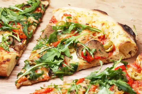 Kawałki pizzy z rukolą, oliwkami i warzywami na drewnianym tle.