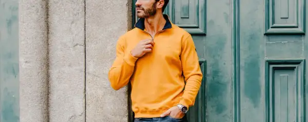 Un homme ethnic blue porte un pull jaune vif avec un col bleu marine, ajustant la fermeture éclair. Il est appuyé contre un mur en pierre.