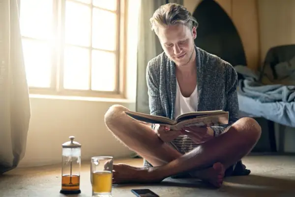 Hombre joven leyendo un libro, disfrutando del **slow living** junto a una ventana con luz natural y una taza de té.