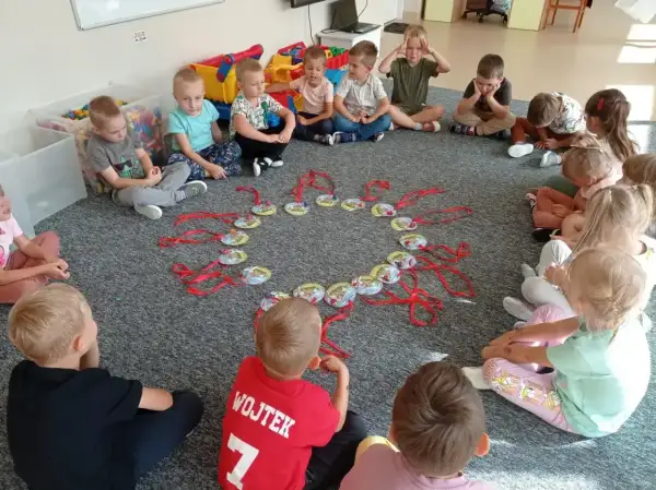 Dzieci siedzą w kółku na dywanie, patrząc na okrąg z zawieszkami. Może to być część zabawy, jak w "Mam chusteczkę haftowaną".