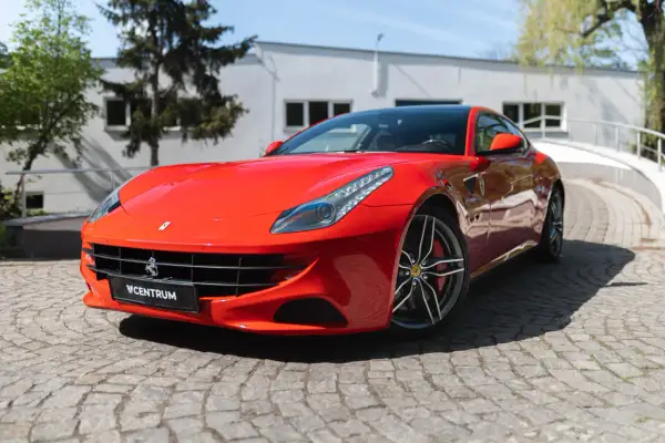Czerwone Ferrari FF na brukowanej drodze, z budynkiem i drzewami w tle.