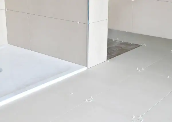 Łazienka w trakcie remontu. Widoczny niski brodzik z białym akcentem i płytki podłogowe z krzyżykami dystansowymi. W tle widać fragment ściany i drzwi.