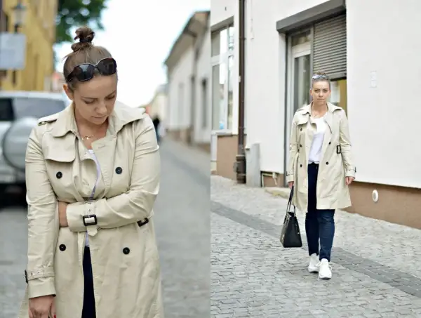 Kobieta w beżowym trenczu i sportowych stylizacjach, z torebką i okularami przeciwsłonecznymi, idzie po brukowanej ulicy.