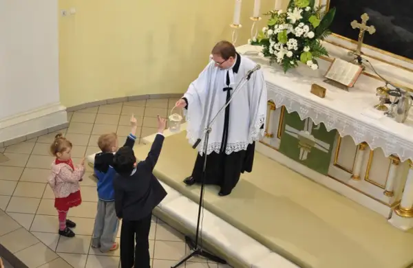 Dzieci z podniesionymi rękami słuchają księdza. Różnice między ewangelikami a katolikami są widoczne w liturgii i symbolach.