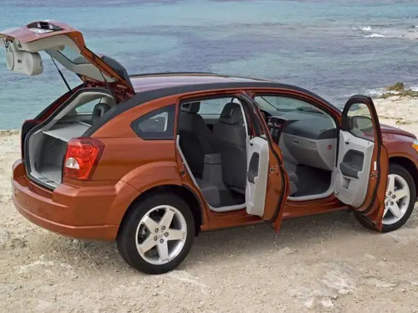 Orange Dodge Caliber 2007 med öppna dörrar och baklucka vid havet. Perfekt för en roadtrip!