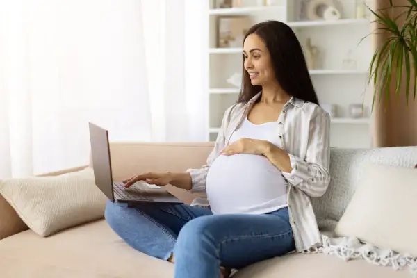 Kobieta w zaawansowanej ciąży sprawdza w internecie informacje o terminie porodu, siedząc na kanapie z laptopem na kolanach.