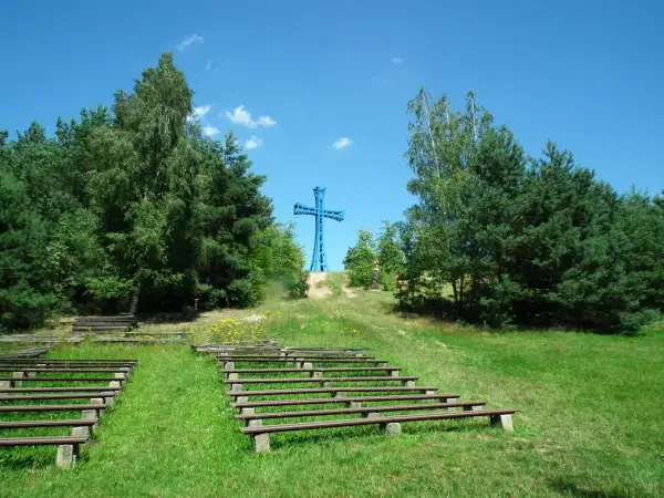 Niebieski krzyż na wzgórzu, najwyższy szczyt wielkopolski, otoczony drzewami. Poniżej amfiteatr z drewnianymi ławami.