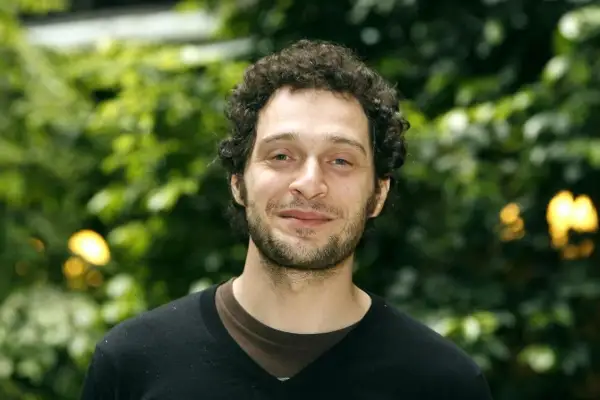 Claudio Santamaria giovane, con capelli ricci e barba, sorride con uno sfondo verde sfocato.