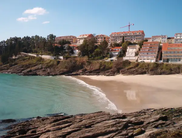 La **playa Caneliñas** se asoma entre rocas y vegetación, con edificios residenciales en la colina y el mar turquesa.