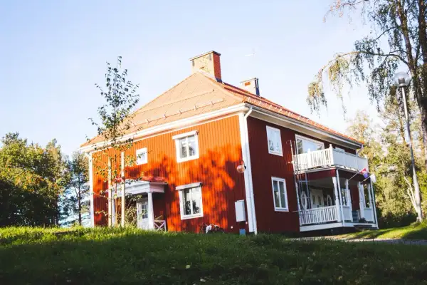 Rött hus med vita knutar och veranda, omgivet av grönska. Ett charmigt vandrarhem i Jokkmokk som bjuder in till en naturnära vistelse.