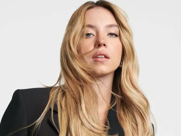 Sydney Sweeney, con su cabello rubio ondulado y un blazer negro, mira directamente a la cámara con una expresión cautivadora.