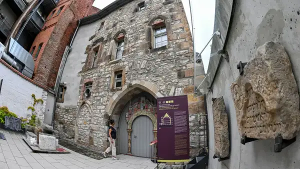 Ein Mann betrachtet die mittelalterliche Steinfassade eines Gebäudes, das Teil des Weltkulturerbes Erfurt ist. Daneben sind alte Steintafeln mit hebräischen Inschriften ausgestellt.